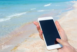 Hand holding a cell phone at the beach in Cancun, Mexico, with clear blue water and white sand in the background.