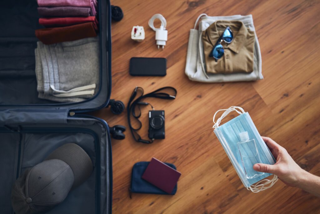 Traveler is preparing for travel in new normal. Hand holding face masks and hand sanitizer for personal protection against packing suitcace with summer clothing, passport and camera.
