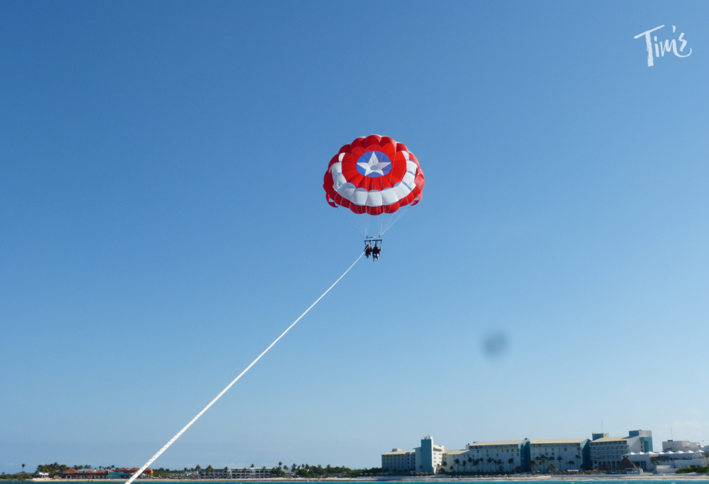 An image of parasailing over the clear turquoise waters of Cancun's ocean. A colorful parasail is seen high above the water, with a person securely harnessed and attached to it, soaring through the sky. The vibrant blue ocean stretches out beneath, reflecting the sunlight. The coastline of Cancun is visible in the distance, with white sandy beaches and lush green palm trees lining the shore. The image captures the exhilarating experience of parasailing while showcasing the natural beauty of the Cancun coastline