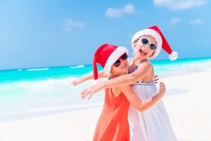 Two smiling girls in Santa hats hugging on the beach in Cancun, enjoying a holiday vacation by the ocean.