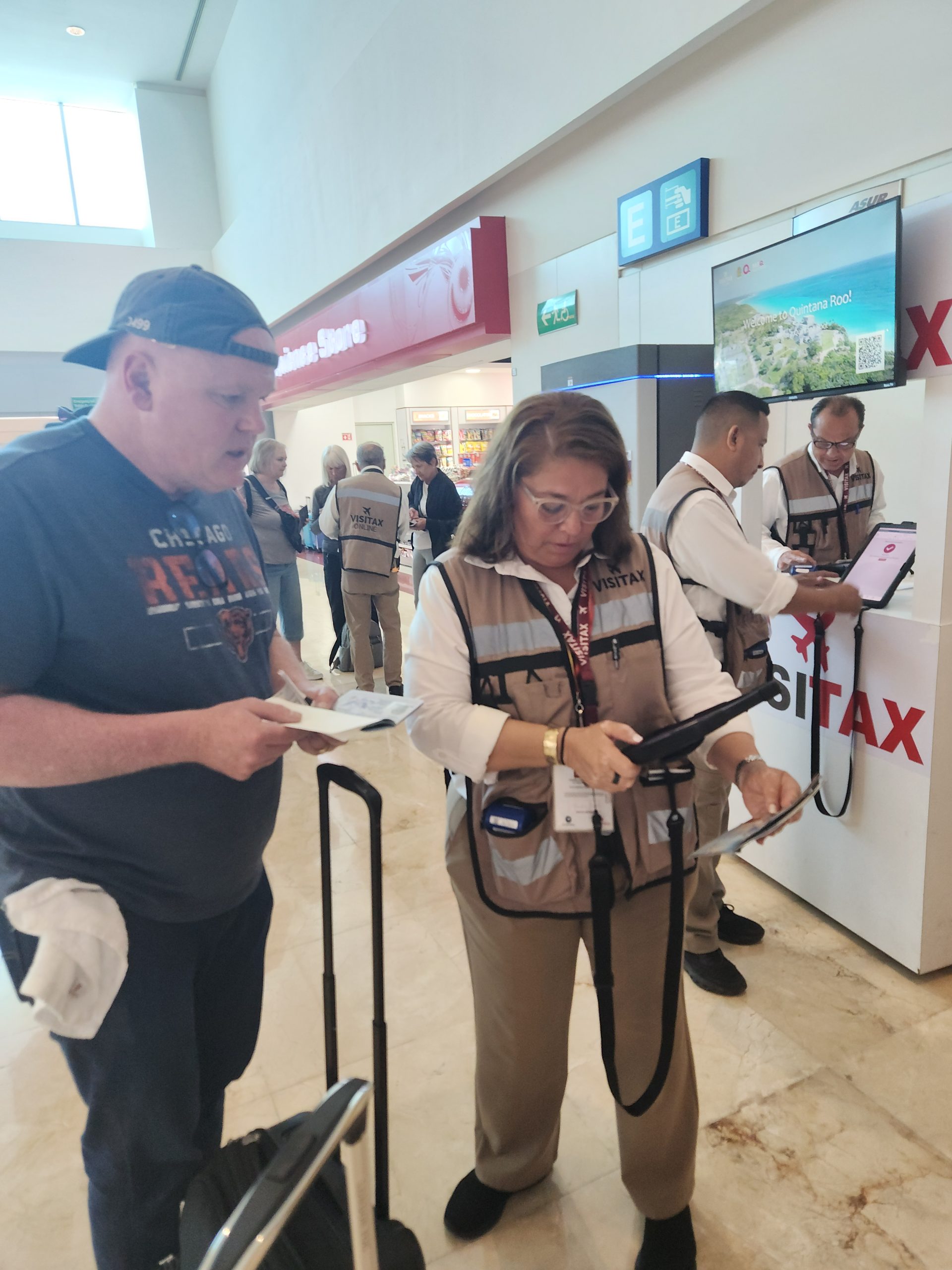 A VISITAX official in Terminal 4 assists Tim with paying the required tourism tax before leaving Cancun in May 2025. Tim stopped by VISITAX official inside Cancun Airport Terminal 4 using a tablet to process payment before departure in May 2025.