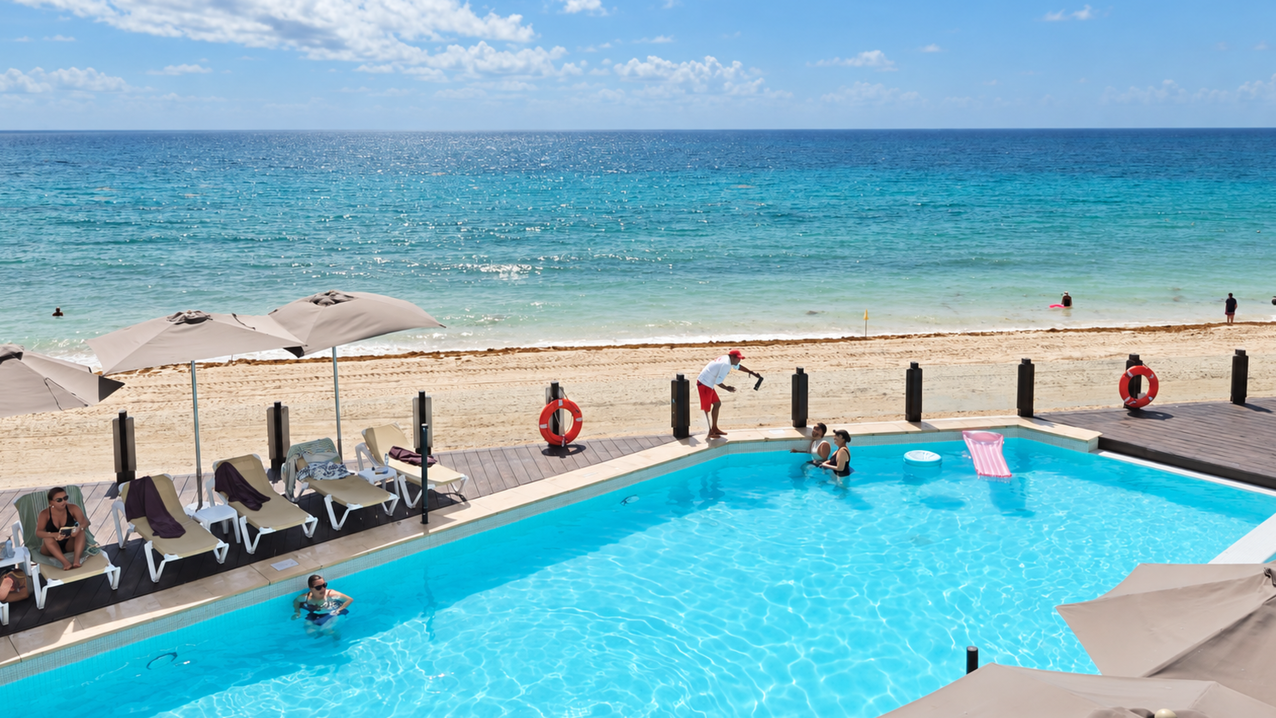 Beachfront swimming pool with swimmers, lounge chairs, and umbrellas beside the Caribbean Sea in Cancun